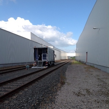 Blick auf Gleise mit einem grauen Zugwagen zwischen zwei Lagerhallen unter blauem Himmel.