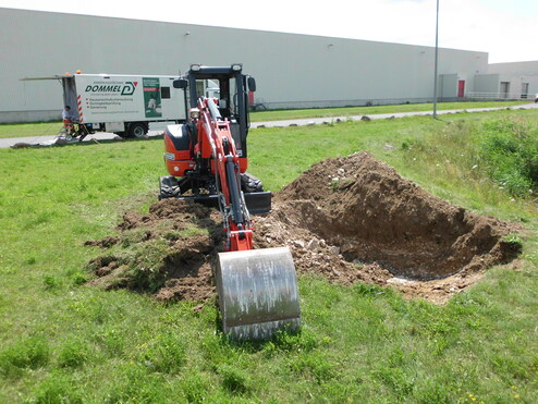 Bagger arbeitet auf einer Wiese, neben einem Lkw mit Aufschrift "Dommel" und einem ausgehobenen Erdloch.