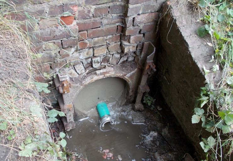 Abfluss&ouml;ffnung mit gr&uuml;nem Schlauch, umgeben von Ziegelmauerwerk und stehendem Wasser im Erdreich.