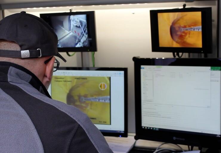 Ein Mann in einer grauen Jacke sitzt vor mehreren Monitoren, die technische Inhalte und Videos zeigen.