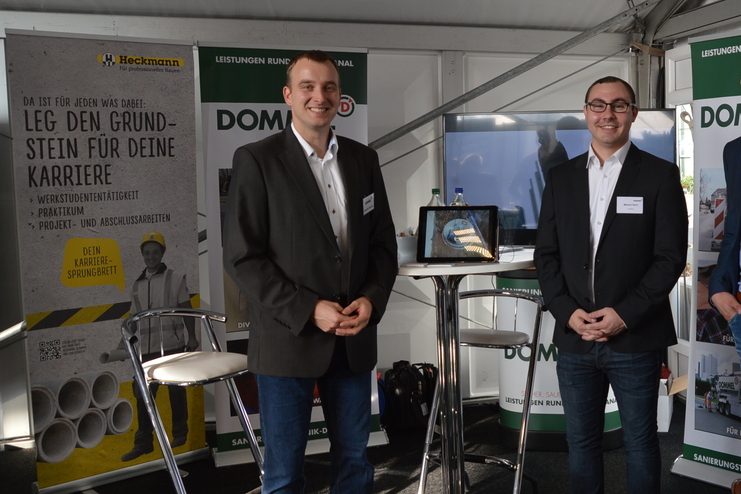 Drei M&auml;nner in Anz&uuml;gen stehen in einem Messezelt vor Karrieretafeln und einem Tisch mit Displays.