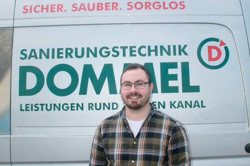 Mann mit Brille steht vor einem Fahrzeug mit der Aufschrift „Sanierungstechnik Dommel“ und einem Slogan.