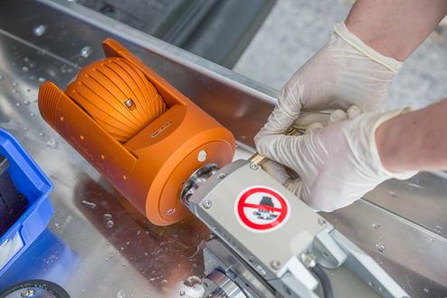 Hände in Handschuhen reparieren ein orangenes Gerät auf einer metallischen Oberfläche mit Wasserflecken.