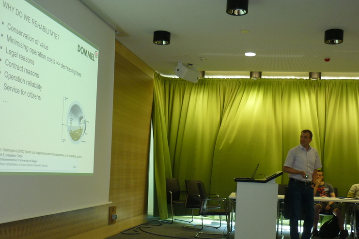 Pr&auml;sentation mit einem Referenten vor einer Tafel, w&auml;hrend Zuh&ouml;rer am Tisch sitzen und Notizen machen.