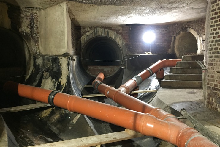 Blick in einen unterirdischen Kanal mit orangefarbenen Rohren, Ziegelwänden und Treppe im Hintergrund.