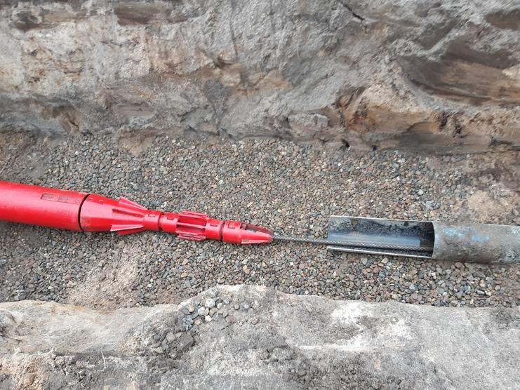 Rotes Bohrwerkzeug liegt neben einem schwarzen Bohrrohr auf einem Schotterboden in einer grabenartigen Struktur.