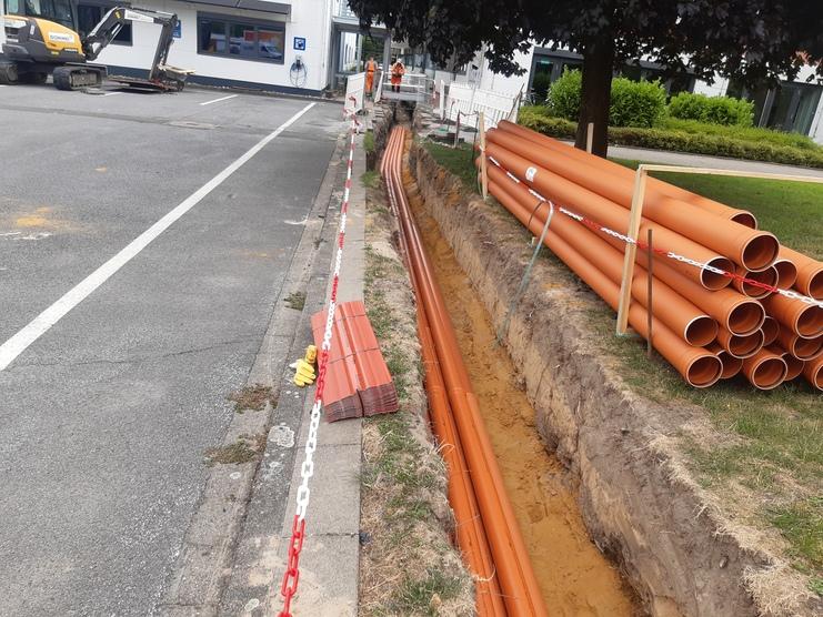 Baufläche mit orangefarbenen Rohren, graben und Absperrungen neben einer Straße und Gebäuden.