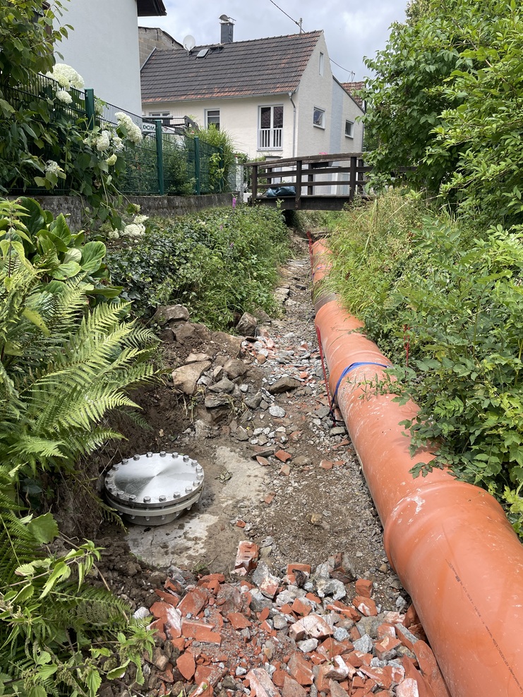 Blick auf eine Baustelle mit orangefarbenem Rohr, Steinresten und einer runden Abdeckung, umgeben von Pflanzen und einem Haus.