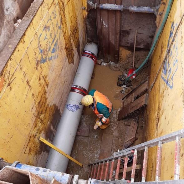 Blick in einen Graben mit Arbeiter, der eine graue Rohrleitung inspiziert, umgeben von nassen, erdigen Wänden.