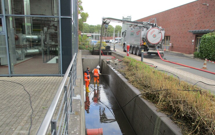 Arbeiter in Schutzkleidung reinigen einen schmalen Wassergraben neben einem Gebäude mit Hochdruckgerät.