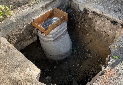Betonrohr in einer offenen Grube, umgeben von beschädigtem Asphalt und Gras. Holzrahmen oben darauf.