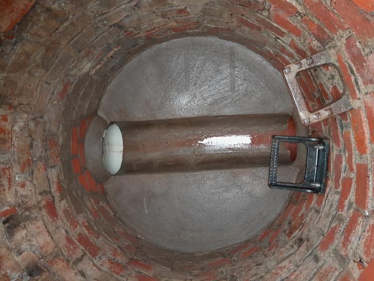 Innenansicht eines runden Betontanks mit Ziegelmauerwerk, einer Metallröhre und zwei Metallteilen am Boden.