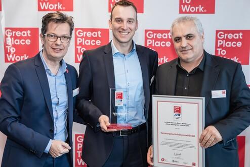 Drei Männer halten Auszeichnungen vor einem Banner mit dem Schriftzug „Great Place To Work 2019“.