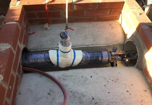 Pipeline mit Anschlussstück in einer Baugrube, umgeben von Ziegelsteinen und rotem Schlauch.