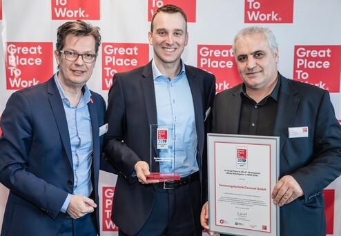 Drei Männer halten Auszeichnungen vor einem Banner mit dem Schriftzug „Great Place To Work 2019“.