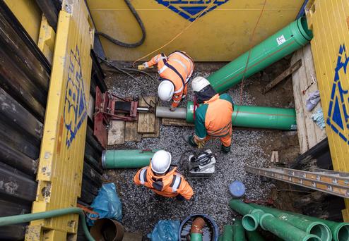 Drei Arbeiter in Schutzkleidung montieren grüne Rohre in einer Baugrube, umgeben von Werkzeugen und Materialien.