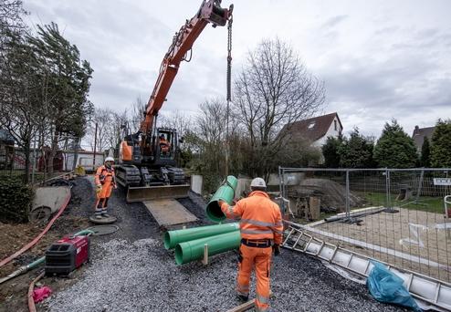 Bauarbeiter in orangefarbener Kleidung heben grüne Rohre mit einem Kran auf einer Baustelle mit Schotteruntergrund.