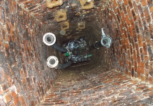 Blick in einen runden, mit Ziegeln gemauerten Schacht mit drei Rohren und Wasser am Boden.