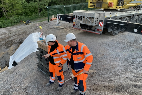 Zwei Bauarbeiter in orangefarbenen Schutzanzügen besprechen sich auf einer Baustelle mit einem Lastwagen im Hintergrund.