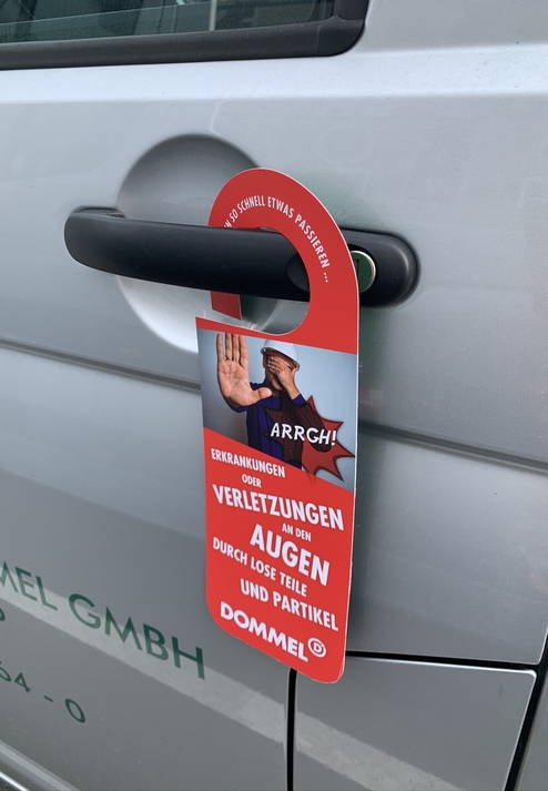 Schilder hängt an einer Autotür und warnt vor Augenverletzungen durch lose Teile.