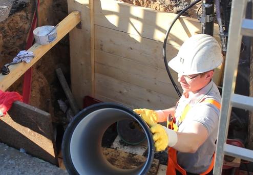 Bauarbeiter mit Helm und Handschuhen hebt ein Rohr aus einer Erdgrube, umgeben von Holzwänden und Werkzeugen.