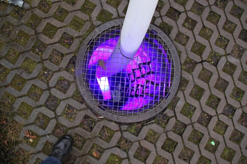 Nahaufnahme eines Gullys mit rötlichem Licht und einem Fuß auf einem gemusterten Gehweg aus Pflastersteinen.