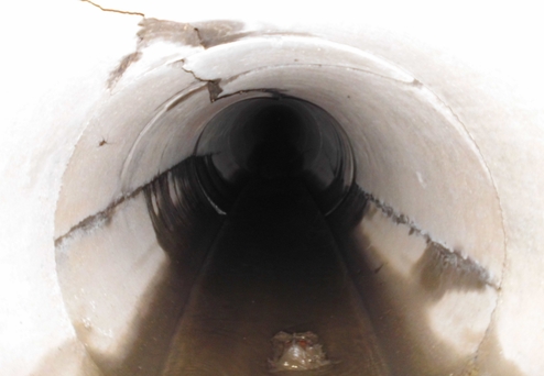 Blick in einen dunklen Abwasserkanal mit feuchtem, heruntergefallenem Schlammboden und sichtbaren Rissen in den Wänden.