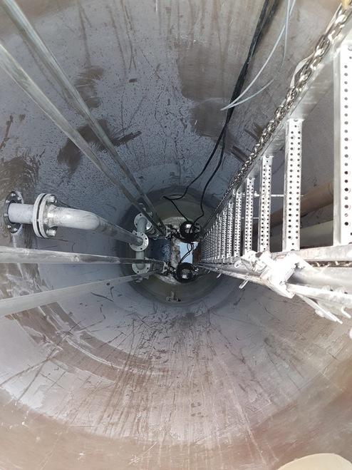 Blick nach unten in einen zylindrischen Schacht mit einer Leiter und Rohrleitungen an der Wand.