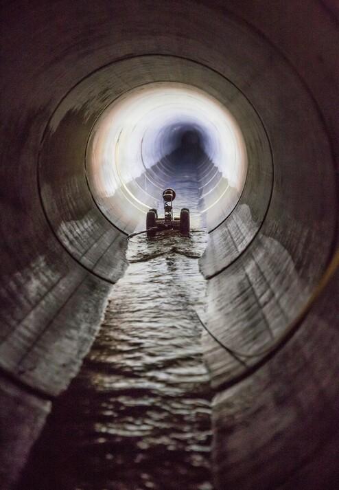 Fernansicht eines fahrzeugähnlichen Objekts in einem dunklen Rohr mit Wasser auf dem Boden und beleuchtetem Hintergrund.