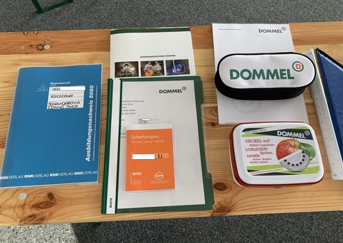 Bürotisch mit Unterlagen, Sicherheitsausweis, Schreibutensilien und einer Plastikdose mit dem Logo von Dommel.