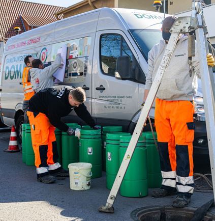Drei Arbeiter in orangefarbener Kleidung bereiten grüne Behälter vor, während ein Fahrzeug im Hintergrund parkt.